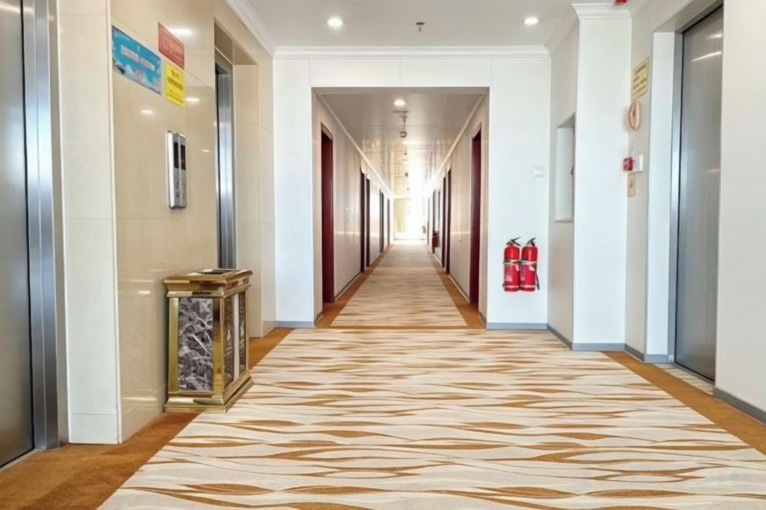 A hotel corridor with a patterned carpet, fire extinguishers, and a fire hose on the right wall.