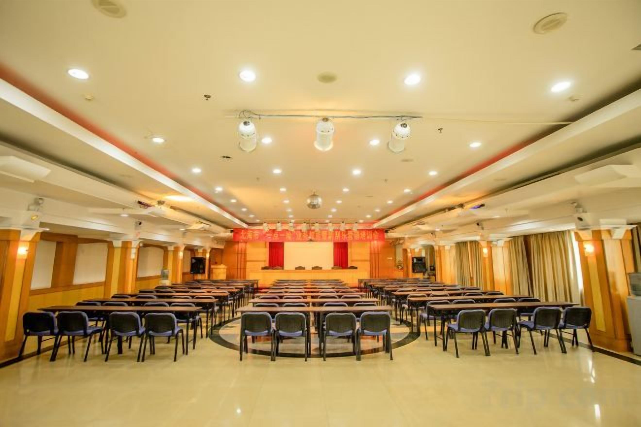 A conference room with rows of chairs, a stage with a red backdrop, and a ceiling with recessed lighting.