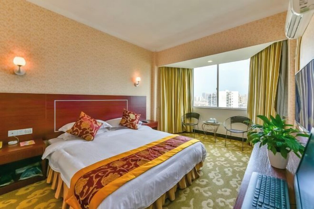Hotelkamer met een bed, een bureau met een laptop, een stoel, een potplant en een raam met gordijnen.