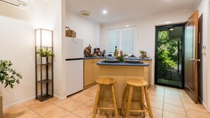A compact kitchen with a built-in fridge, a window with blinds, two stools, and a door leading to a garden.