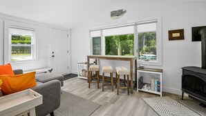 Una sala de estar moderna con un sofá gris, una mesa de comedor de madera con cuatro sillas, una chimenea y ventanas grandes que ofrecen vistas a la vegetación.