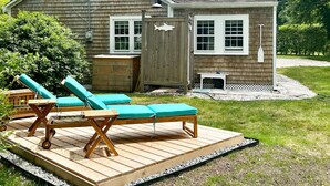 Una terraza de madera con dos sillones de descanso y una pequeña mesa, un cobertizo de madera, y una casa con techo de tejas y molduras blancas.