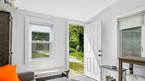 Un interior luminoso y blanco con una puerta que da a un jardín, un sofá gris con un cojín naranja y una pequeña mesa con libros.