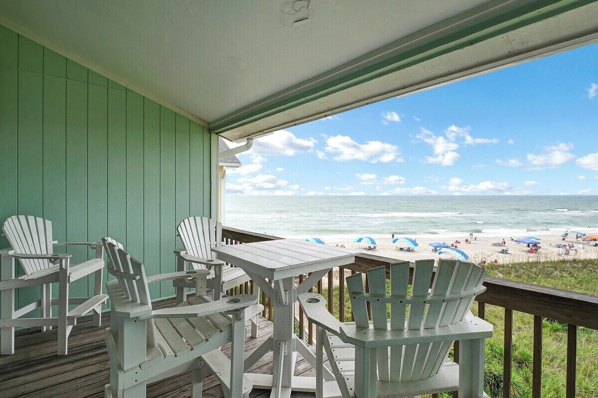 A covered deck with Adirondack chairs and a table, overlooking a beach with umbrellas and people.
