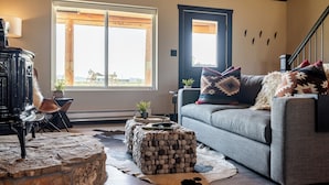 A cozy living room with a stone fireplace, a gray sofa with patterned cushions, and a large window with wooden beams. - Cedar Mountain Retreat- Hot Tub, Pet Friendly, Mountain Views (Cody)