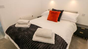 Ein ordentlich gemachtes Bett mit einer schwarz-weißen Steudecke, zwei Kissen und einem roten Zierkissen. Zwei Nachttische mit Lampen und ein Heizkörper unter dem Bett.