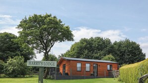 Una cabana rústica amb un rètol que diu "Harry's Lodge", envoltada de vegetació abundant i una paret de pedra.