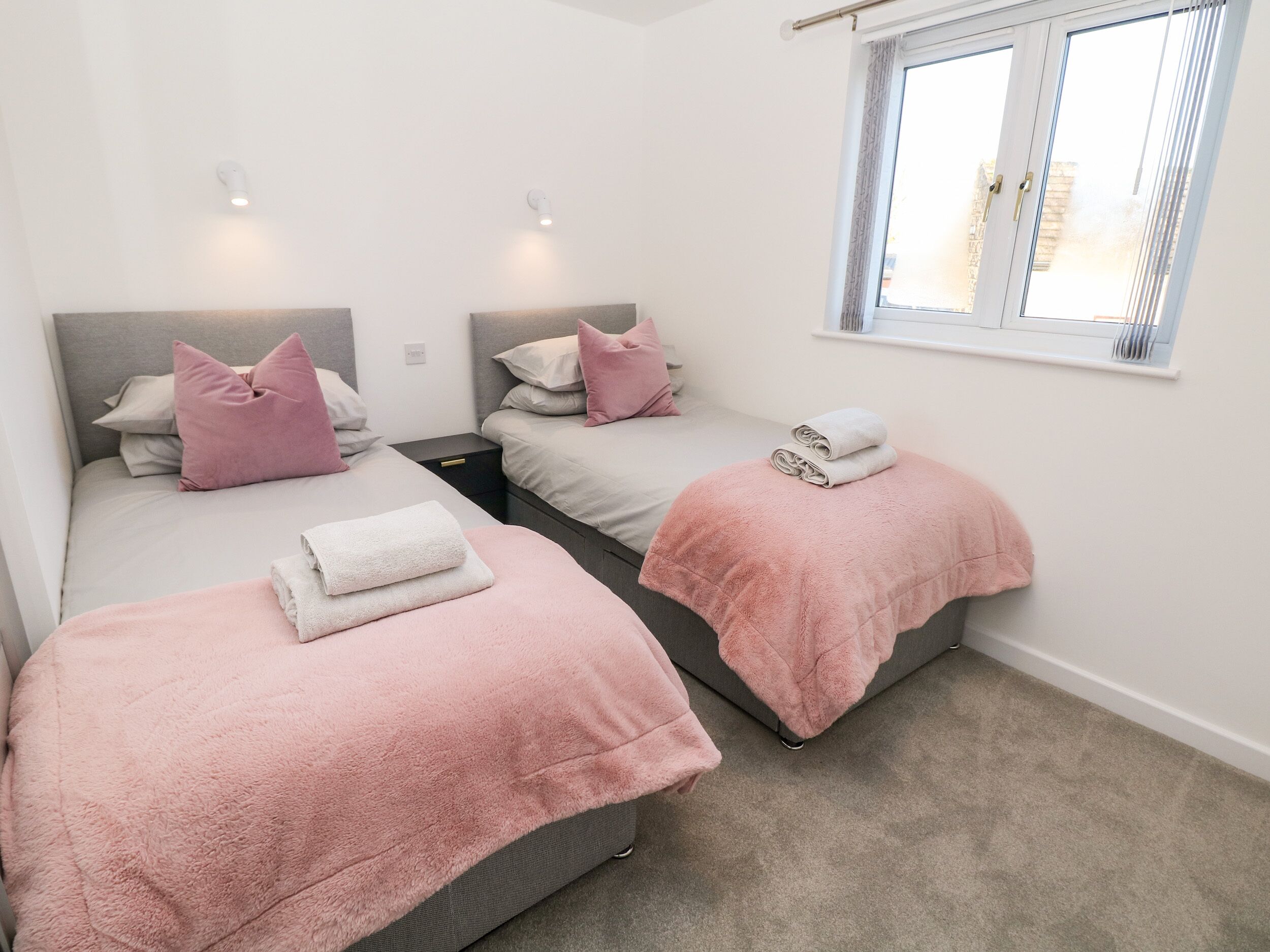 A bedroom with two beds, each with a pink blanket and a white towel. There are bedside tables and a window with curtains.