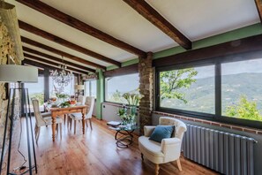 Comedor con mesa de madera, sillas y una araña. Se ve el exterior a través de amplios ventanales.