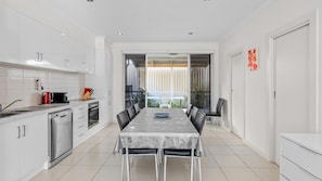 A modern kitchen with white cabinetry, a dining area with a table and chairs, and a view of a balcony with plants. - No.8 - The Block (Victor Harbor)