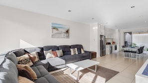 A modern living room with a grey sectional sofa, a white coffee table, and a kitchen area with white cabinets and stainless steel appliances. - No.8 - The Block (Victor Harbor)
