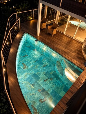 Una piscina con muelle de madera y paredes de vidrio, rodeada de mobiliario exterior.
