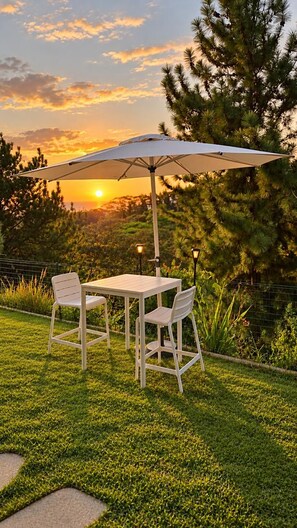 Un área de estar al aire libre con una mesa y sillas bajo un gran paraguas, rodeada de follaje y un atardecer de fondo.