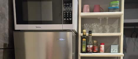 Un refrigerador de acero inoxidable con un microondas encima, una estantería blanca con cristalería, botellas y un tostador.