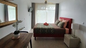 A bedroom with a bed, a bedside table with a lamp, a wooden desk with a chair, a mirror, and a window with curtains.
