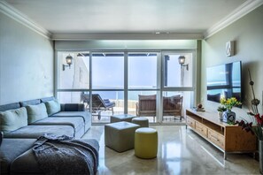 A modern living room with a sofa, ottomans, a TV, and a balcony with a view. - MAMAD - Netanya Panoramic Sea-View 4BR Apartment (Netanya)
