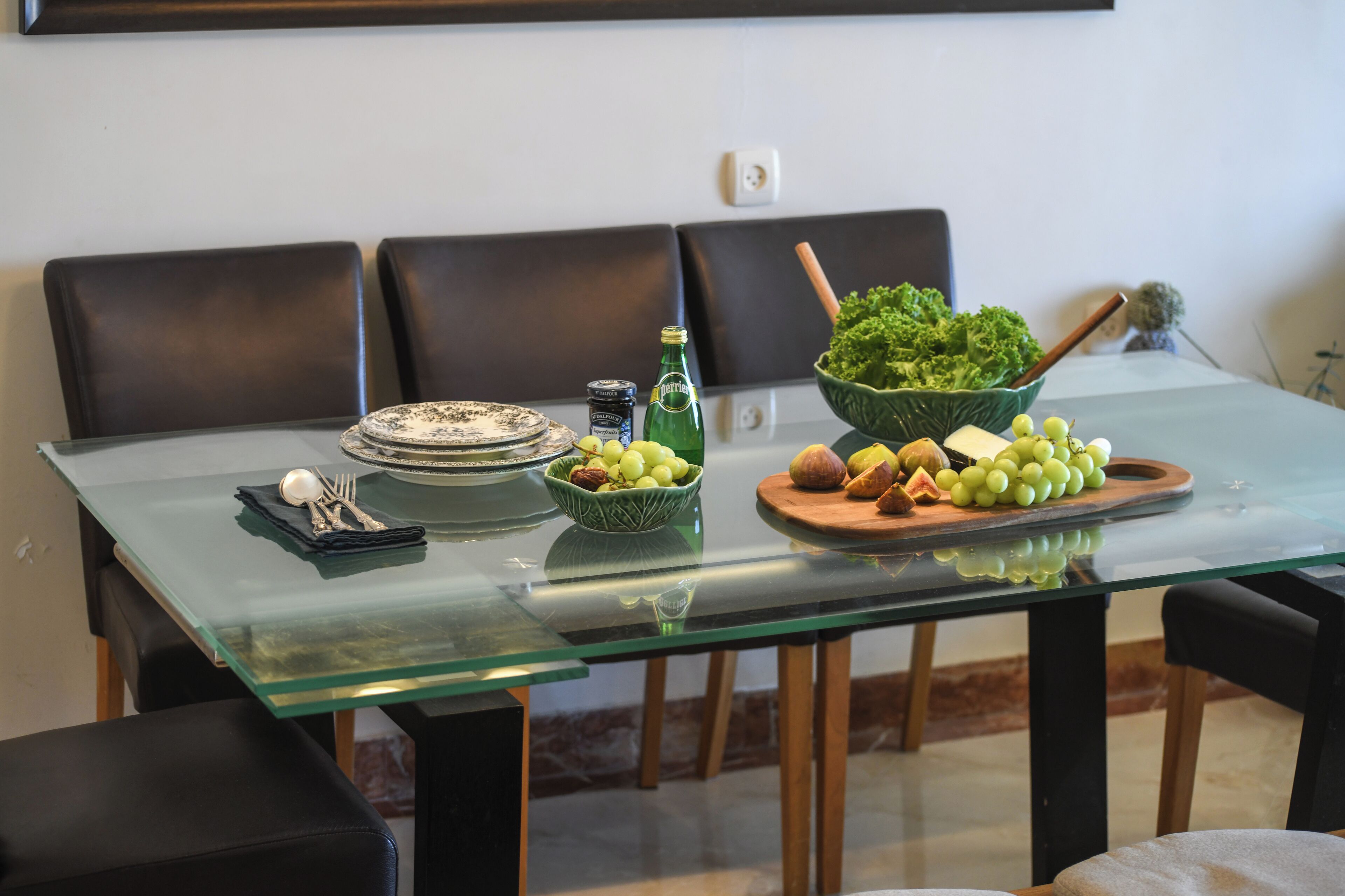 Uma mesa de jantar com tampo de vidro, rodeada por cadeiras pretas. Há pratos, uma tigela de uvas, uma garrafa e uma saladeira.