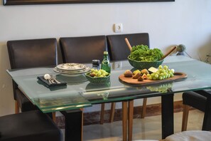 Ein Esstisch mit Glasplatte, umgeben von schwarzen Stühlen. Auf dem Tisch stehen Teller, eine Schale mit Trauben, eine Flasche und eine Schüssel mit Salaten.