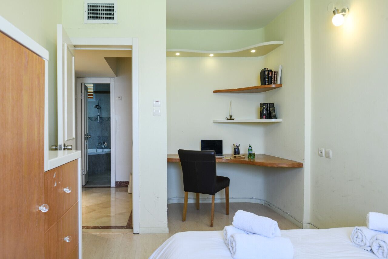 A hotel room with a bed, a desk with a chair, a shower area, and shelves with books and toiletries.