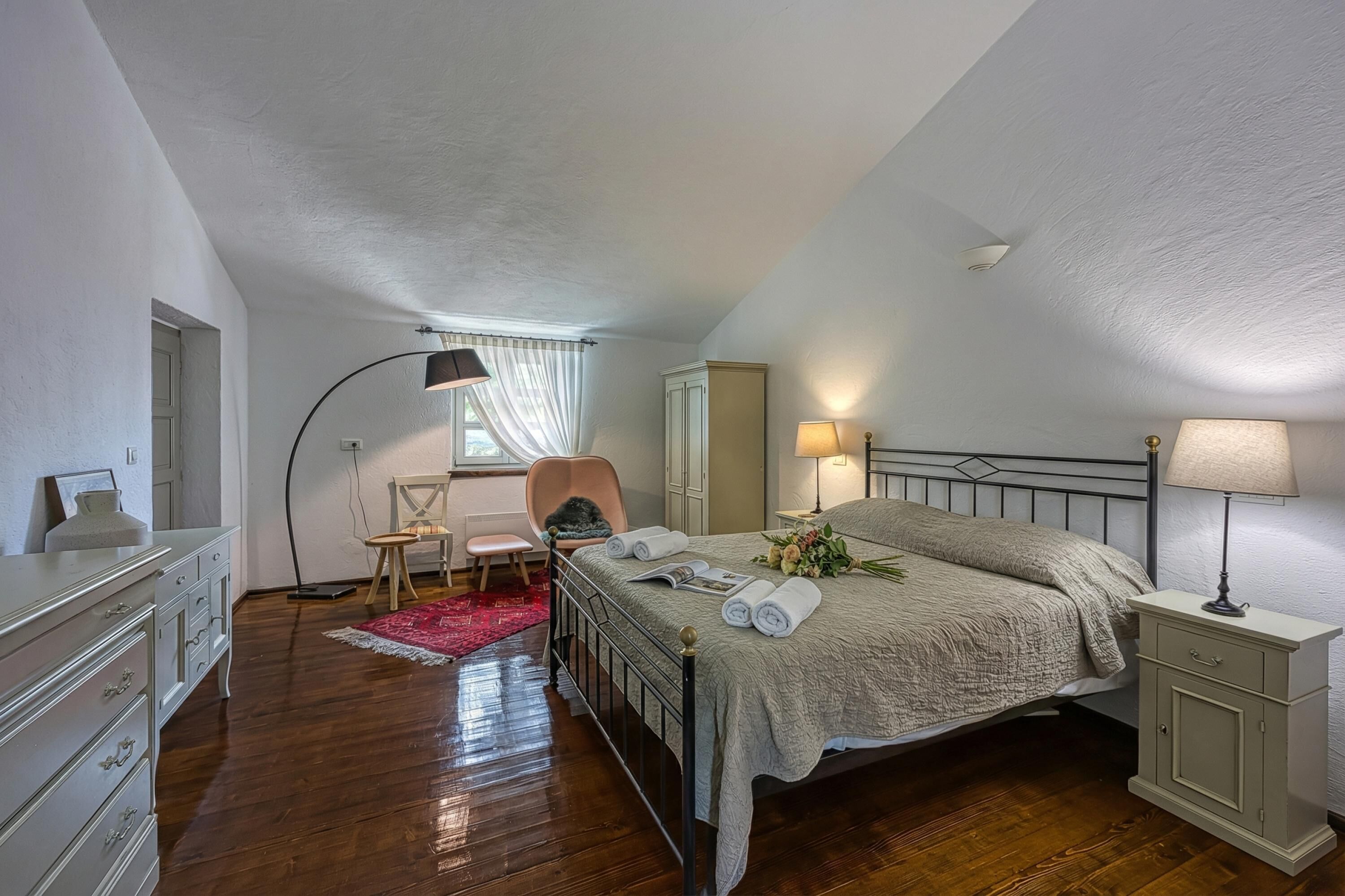 A bedroom with a bed, bedside tables, a chair, a desk, a wardrobe, and a window with curtains.