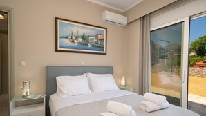 Una habitación de hotel con una cama grande, un cuadro de una escena costera, una puerta de cristal que da a un balcón y una mesita de noche con lámpara.