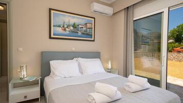 Una habitación de hotel con una cama grande, un cuadro de una escena costera, una puerta de cristal que da a un balcón y una mesita de noche con lámpara.