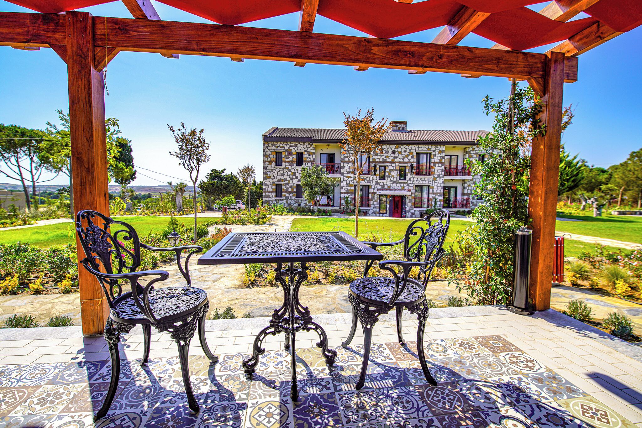 An outdoor patio with a table and chairs, a pergola, and a view of a stone building with a garden in the background.