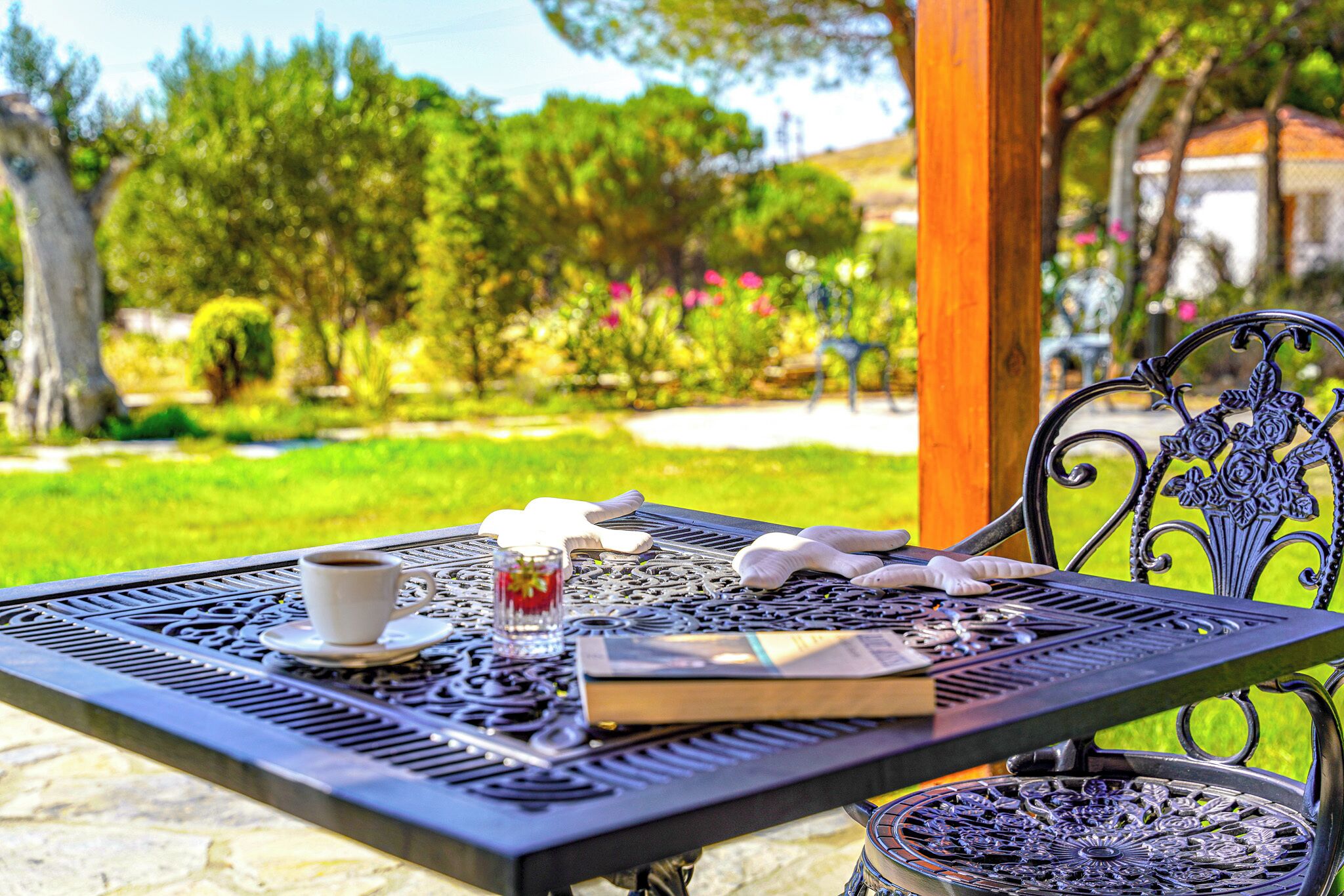A metal table with a cup, saucer, and book, set outdoors with a garden view.