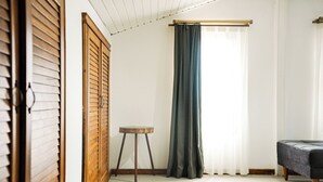 Une pièce avec une armoire en bois, une petite table ronde, un rideau foncé et un lit recouvert d’une couverture grise.