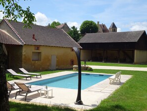 A swimming pool with lounge chairs and a small table, surrounded by a grassy lawn and a building with a tiled roof.