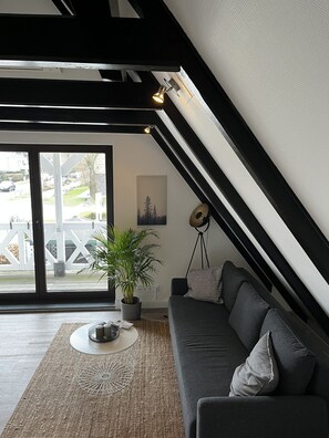 Ein modernes Wohnzimmer mit einem dunkelgrauen Sofa, einem runden Couchtisch und einer Topfpflanze. Ein großes Fenster bietet Blick auf eine Wohngegend.