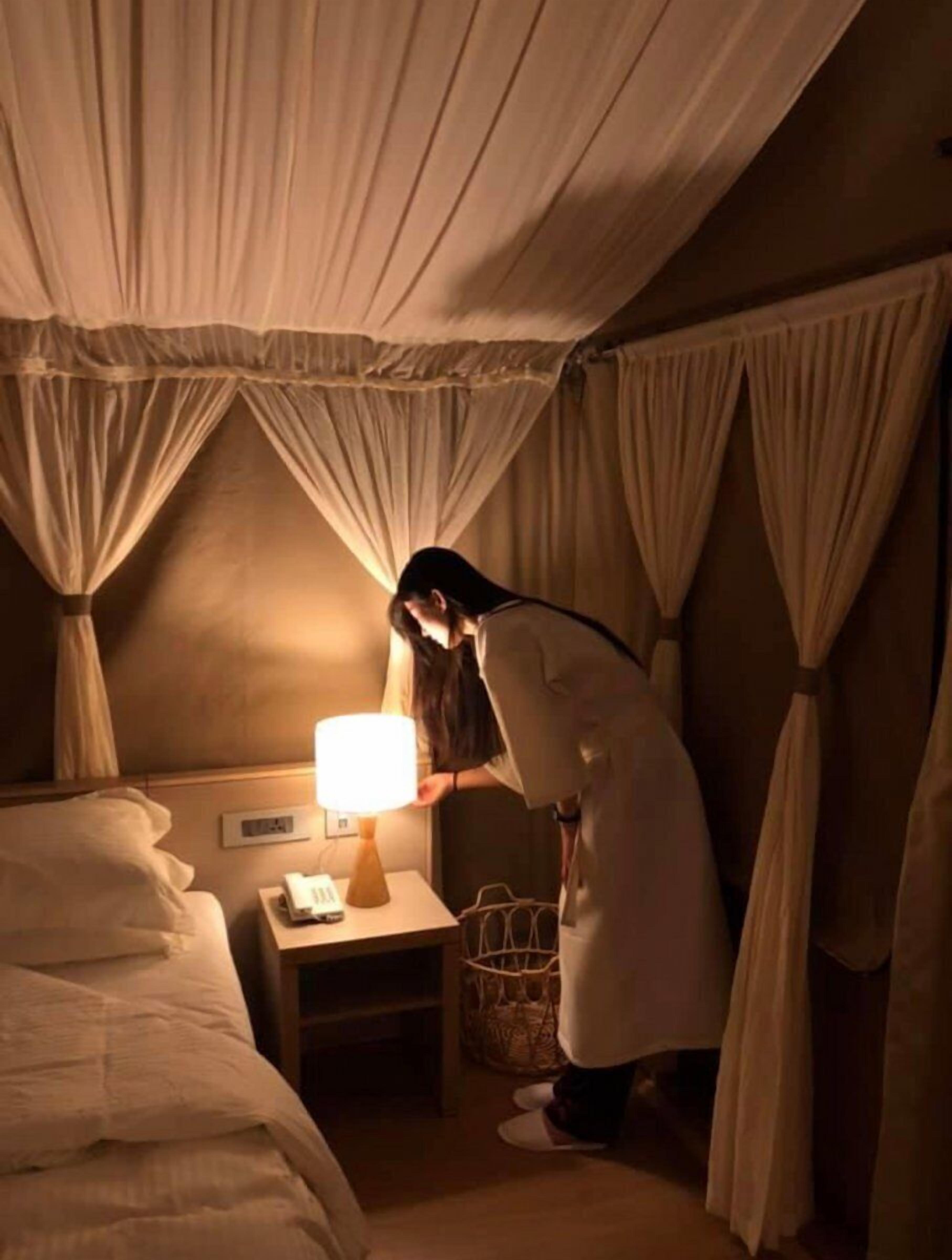 A hotel room with a bed, a bedside table with a lamp, a phone, and a basket.