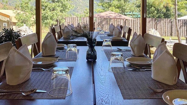 A dining area with tables set for six, large windows offering mountain views, and a wooden deck outside. - The Tiger Nest Camp (Paro)