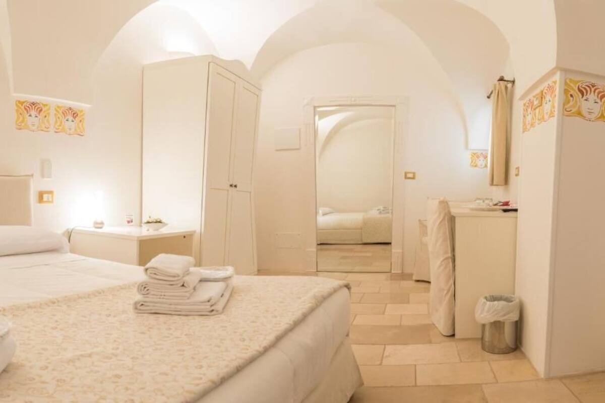 Habitación de hotel con una cama grande, una mesita de noche con lámpara, un espejo y un baño visible a través de un arco.