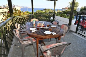 Une table ronde en bois, dressée pour quatre personnes, avec des chaises, une tasse de thé et une assiette de nourriture, sur un balcon donnant sur la mer.