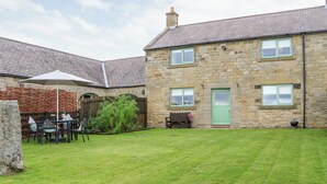 Un bâtiment en pierre avec une porte verte, un jardin clôturé et une terrasse aménagée avec du mobilier d’extérieur.