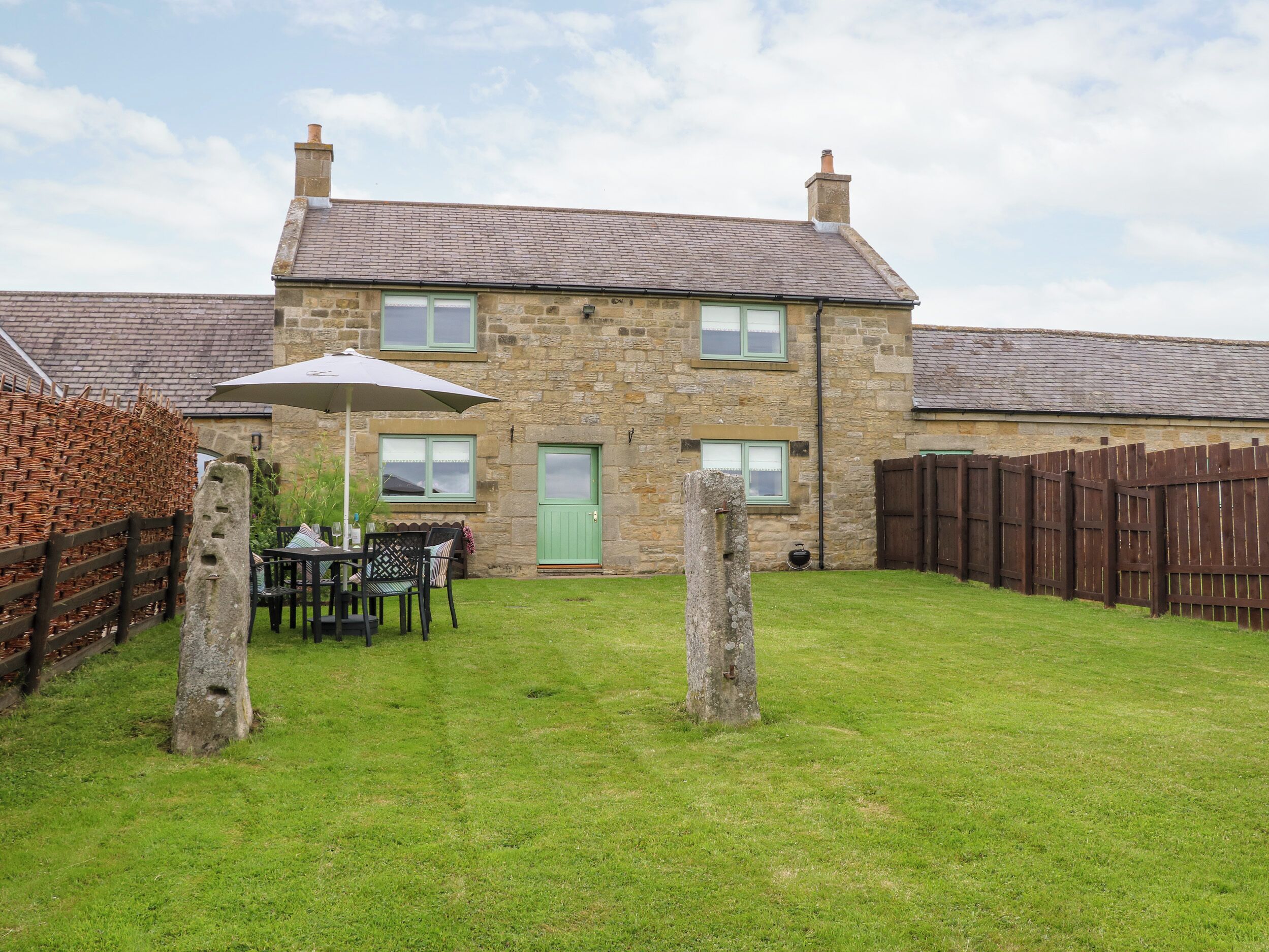 A stone house with a green door, a wooden fence, and a grassy yard with a table and chairs under an umbrella.