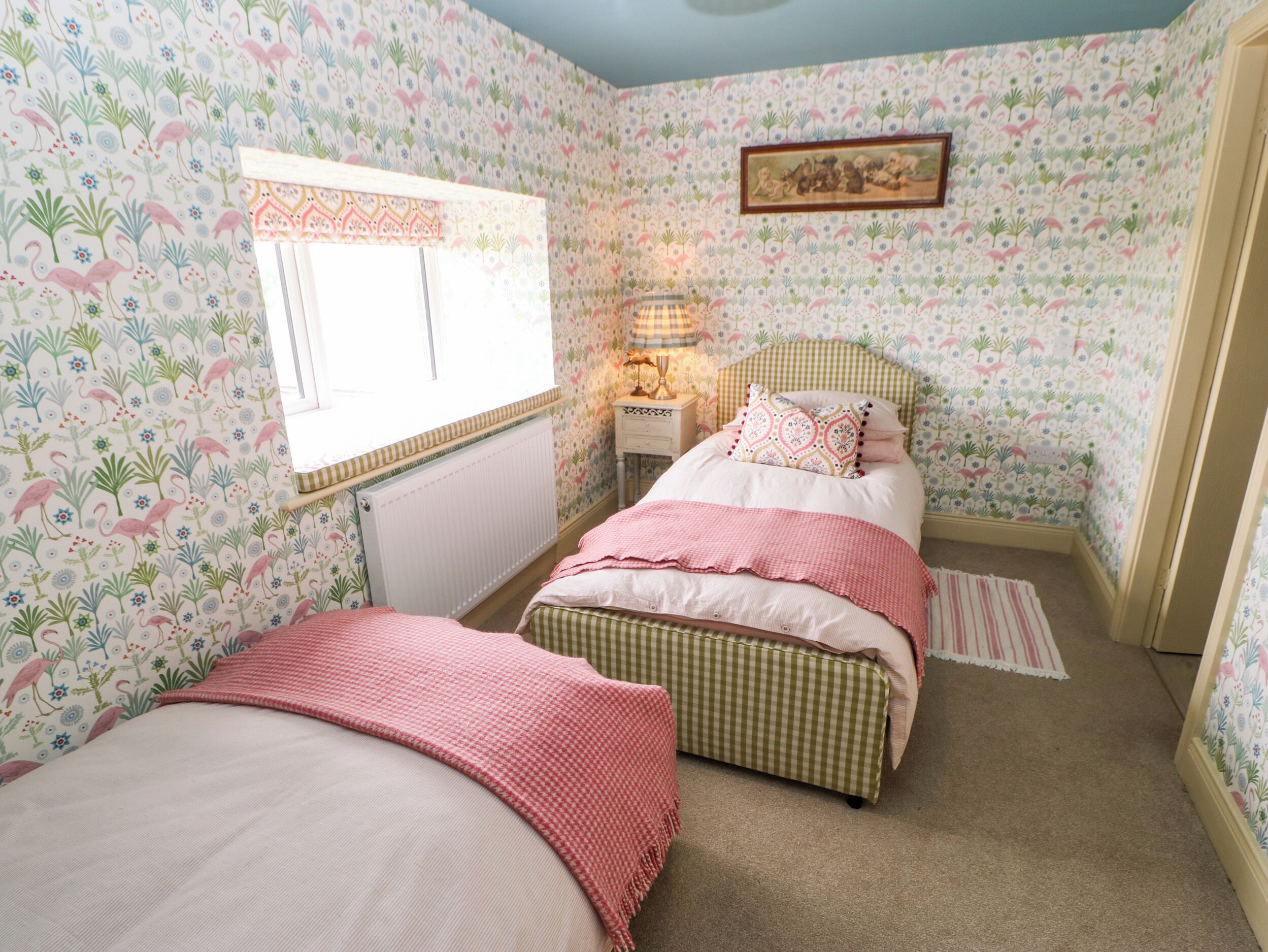 A bedroom with floral wallpaper, two beds with pink bedding, a window, a nightstand with a lamp, and a framed picture on the wall.