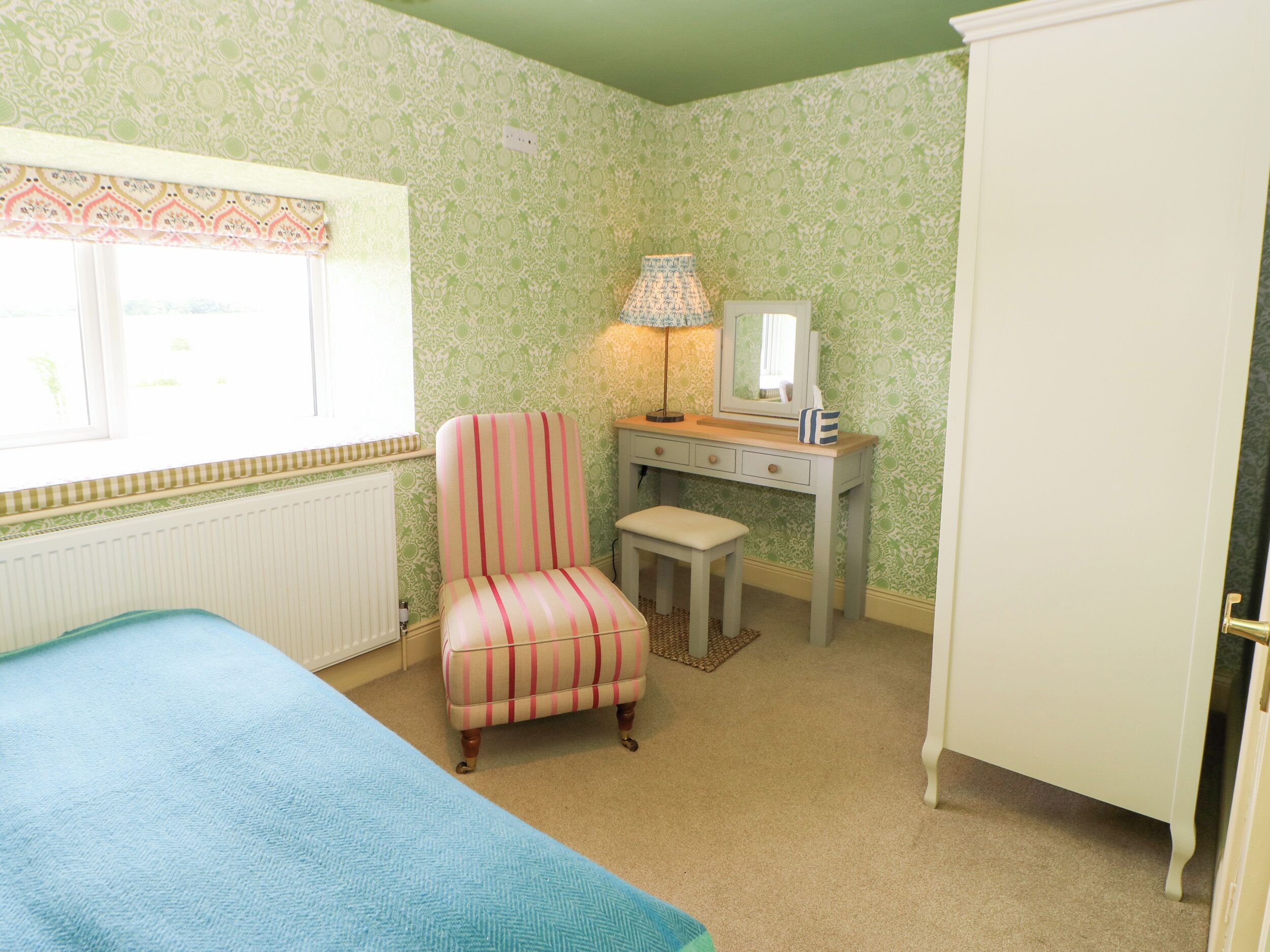 A bedroom with a bed, a chair, a desk with a lamp, a mirror, and a wardrobe.