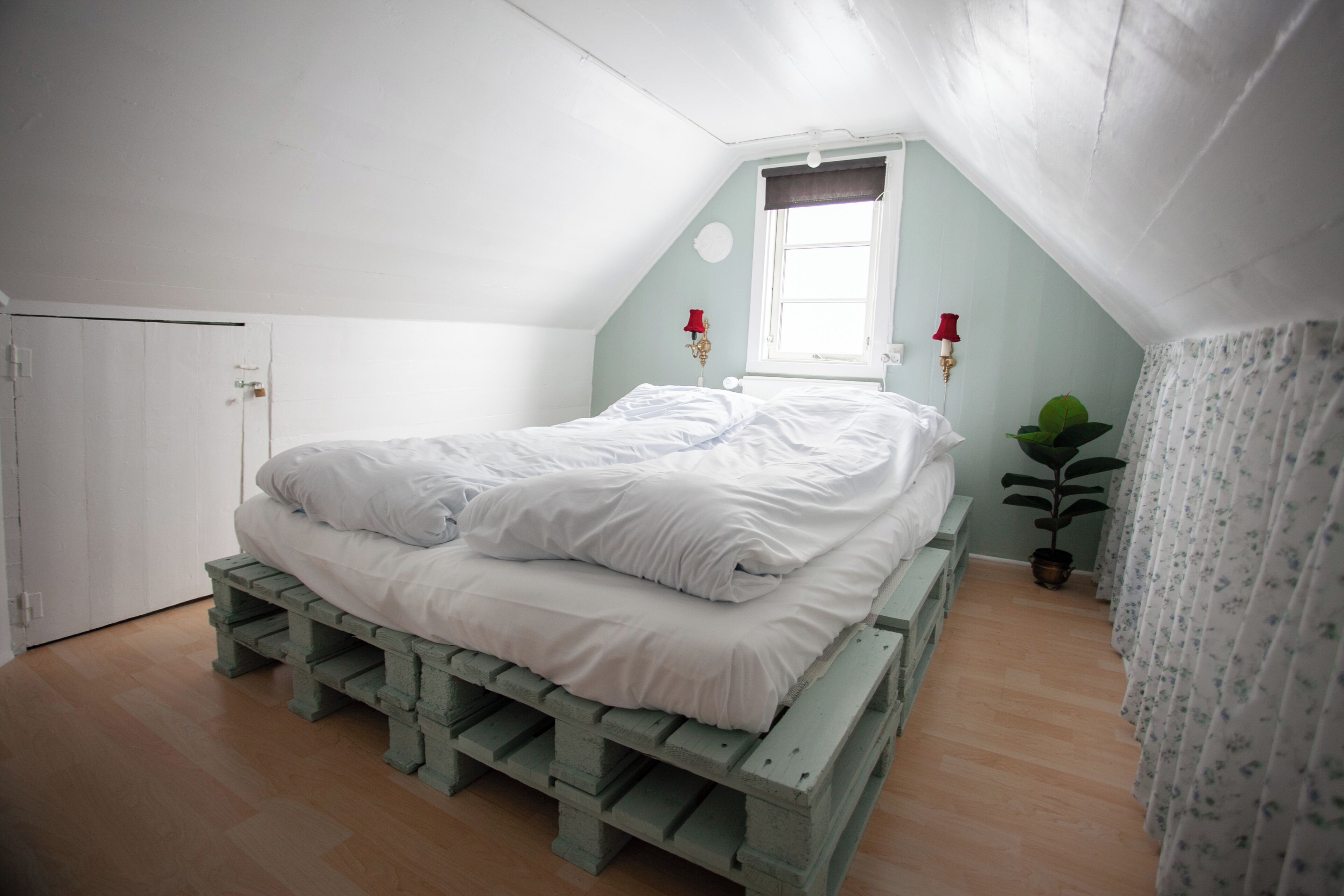 A bedroom with a bed on a wooden pallet, a window with curtains, a potted plant, and a door.