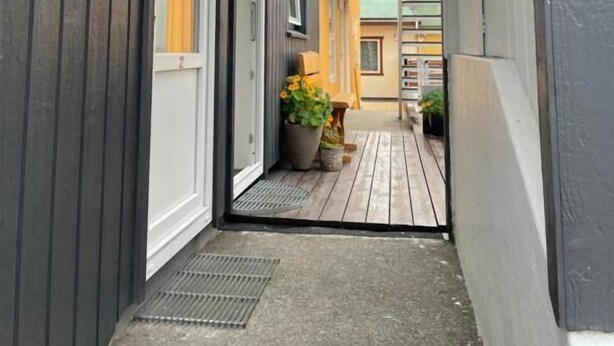 Un couloir étroit au plancher de bois, une grille métallique à l’entrée et une vue sur les bâtiments adjacents dotés de balcons.