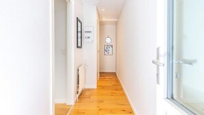 Un couloir étroit avec un plancher en bois et des murs blancs. Des tableaux encadrés ornent les murs, et une porte avec un panneau de verre se trouve sur le côté droit.