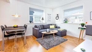 Un salon moderne avec un canapé gris, une table basse ronde et une table à manger avec des chaises. De grandes fenêtres laissent entrer la lumière naturelle et offrent une vue sur la ville.