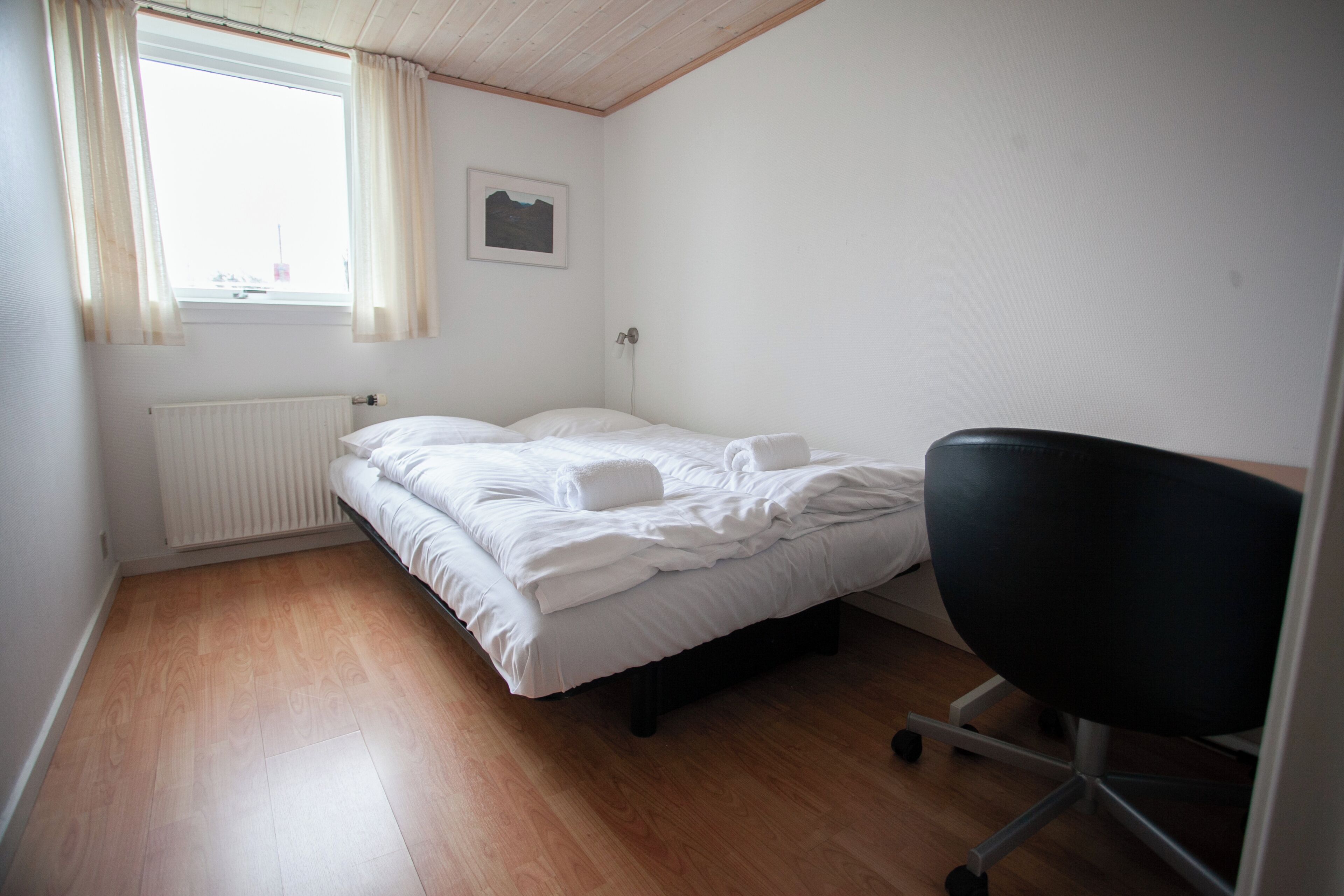 A small, clean room with a single bed, a chair, a radiator, and a window with curtains.