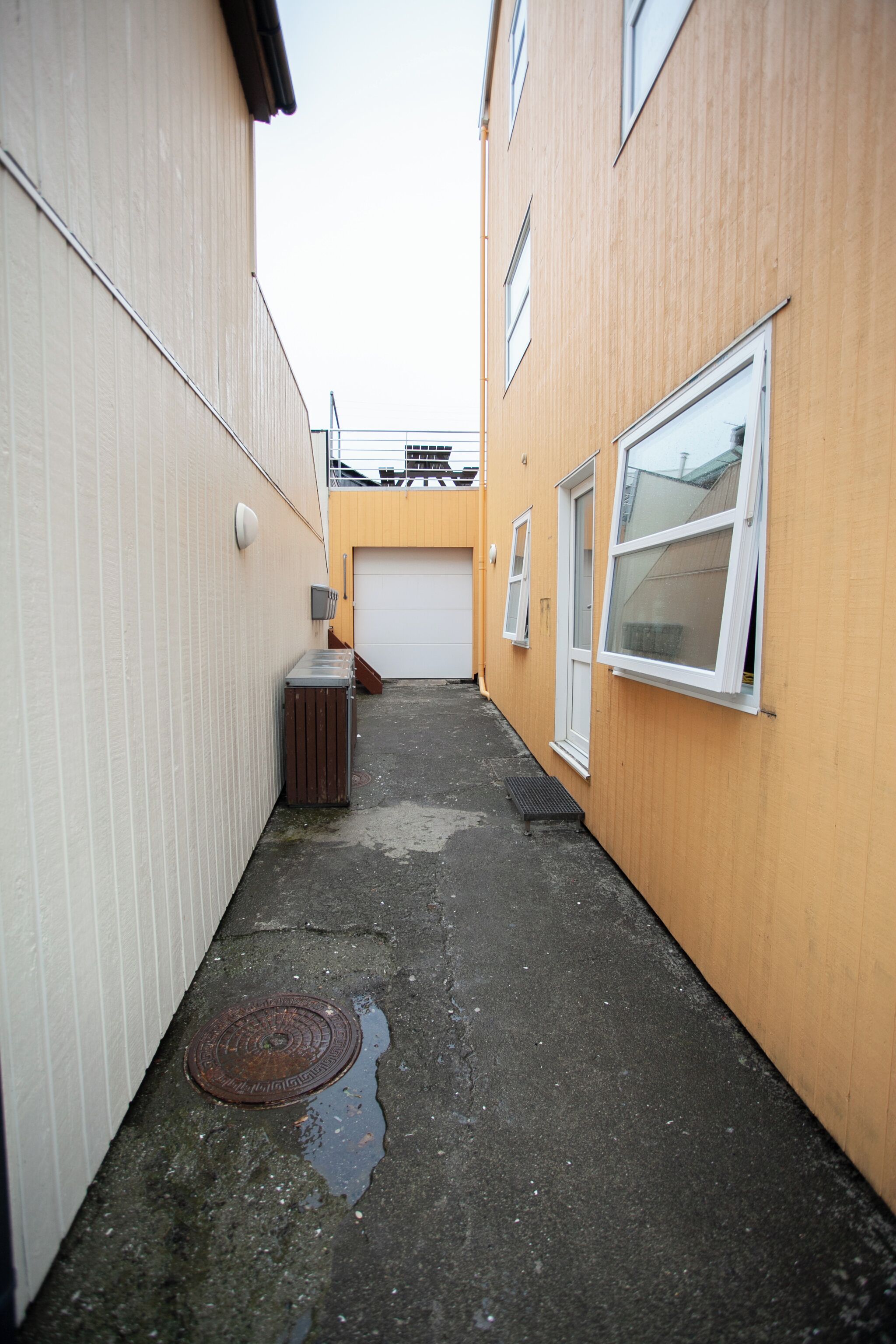 Une ruelle étroite avec un regard d’égout, une poubelle et une porte de garage.