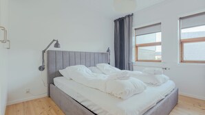 A bedroom with a grey upholstered bed, white bedding, a bedside lamp, a window with curtains, and a wooden floor.
