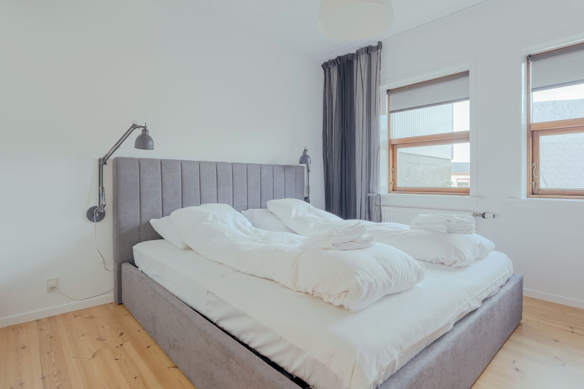 Ein Schlafzimmer mit grauem Polsterbett, weißer Bettwäsche, einer Nachttischlampe, einem Fenster mit Vorhängen und Holzböden.