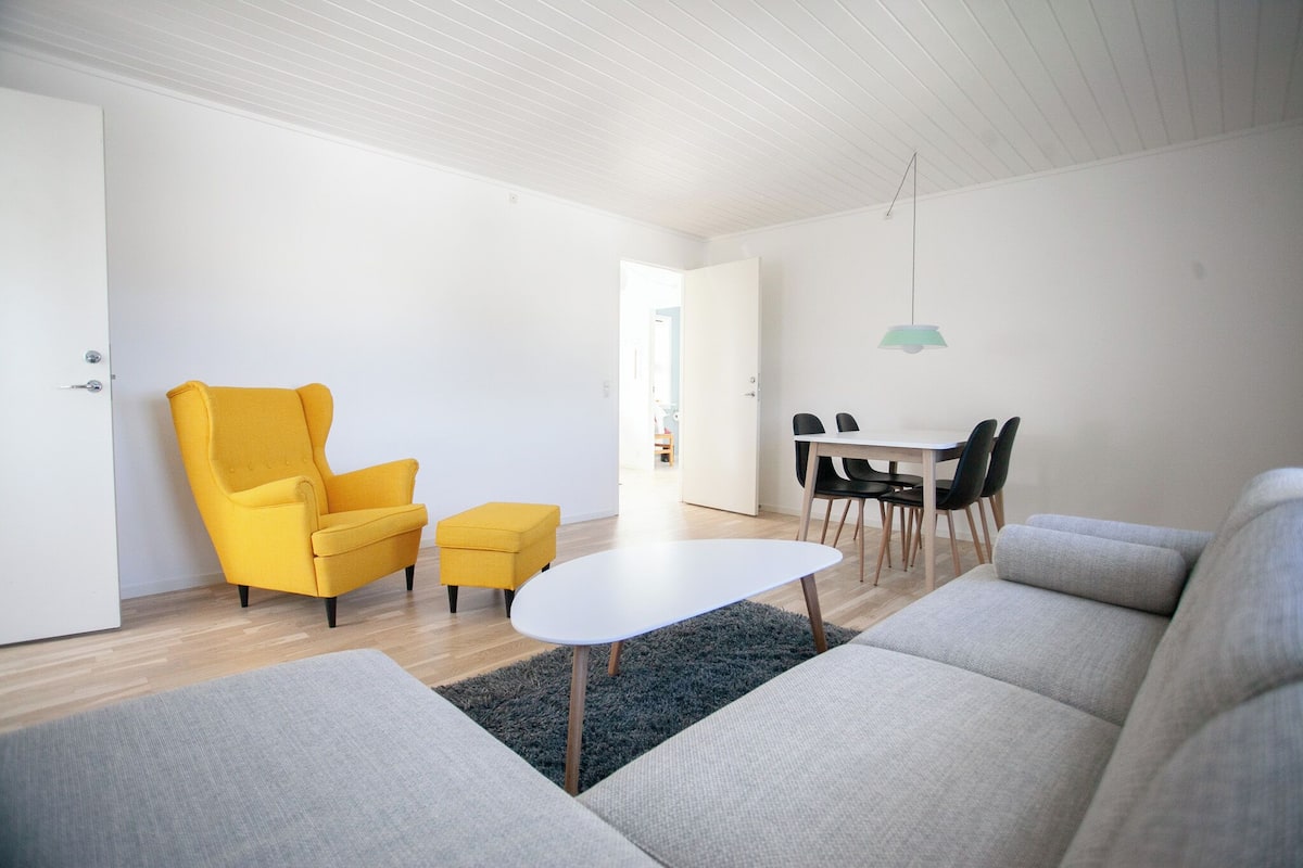 Ein modernes Wohnzimmer mit einer Couch, einem gelben Sessel, einem kleinen Hocker, einem weißen Couchtisch und einem Essbereich mit Tisch und Stühlen.