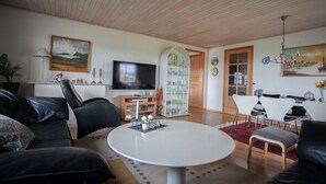 Un salon moderne avec une table basse ronde blanche, des canapés en cuir noir, un téléviseur à écran plat et un coin repas avec des chaises blanches et un tapis rouge.