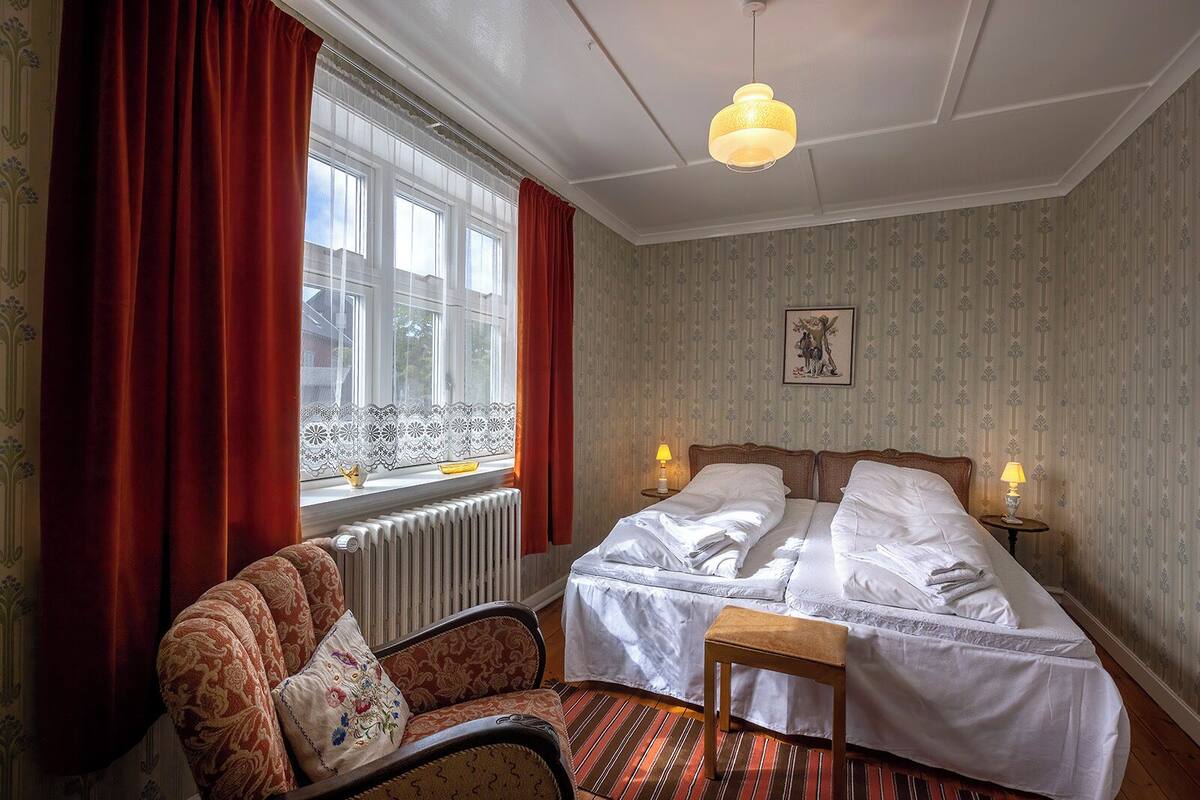 A hotel room with a bed, a chair, a table, a radiator, and a window with curtains.